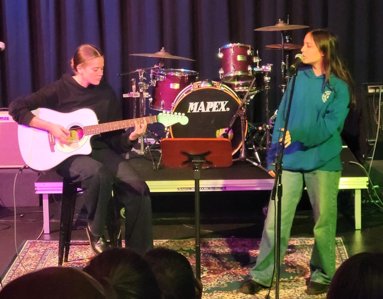 One student singing, and another playing guitar as they perform in the school hall